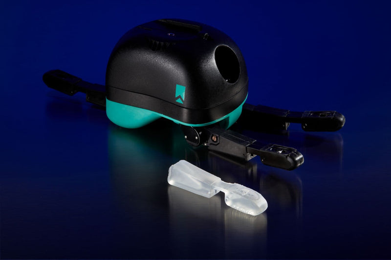 A black and teal drone shown on a dark blue reflective surface, with a small clear plastic accessory placed in front of it.